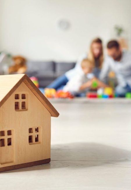 Close-up of miniature detached house on floor at home with happy family playing with little child in background. Mortgage, real estate business, buying property and confidence about future concepts